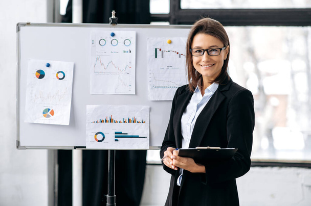 Portrait positive pretty, elegant caucasian business woman with glasses, financial manager or mentor, broker, marketer, stand in the office near whiteboard with graphs, looks at camera, smile friendly - Foto, Bild