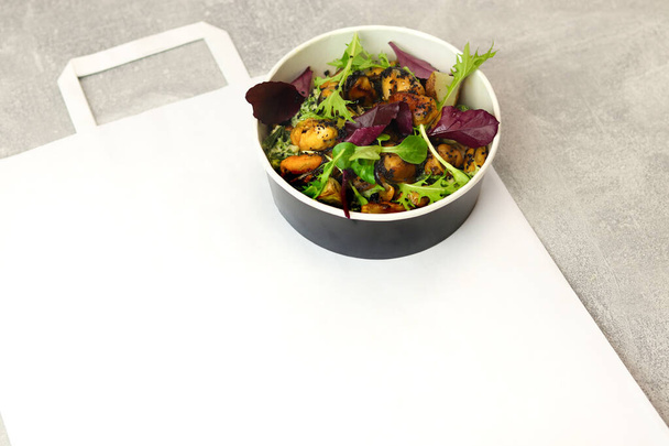 Salad with mussels, arugula and greens in a round takeaway paper container on gray background. Healthy food delivery concept. Banner. Top view. Free space for text. - Foto, Imagen