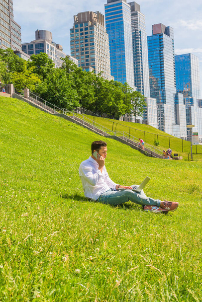 Путь к успеху. Носит белую рубашку, джинсы, кроссовки, молодой студент колледжа сидит на зеленой лужайке с лестницей в деловой район в Нью-Йорке, работает за ноутбуком, разговаривает по телефону - Фото, изображение