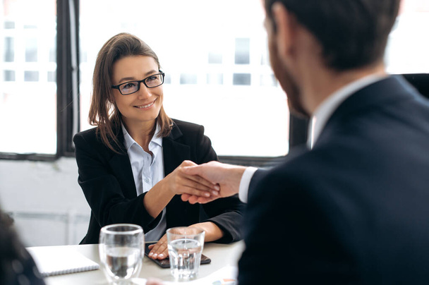Making a deal, shaking hands, confirms agreements. Caucasian intelligent successful business woman, top manager, ceo company, shakes hands with male colleague in modern office, smiling at each other - 写真・画像