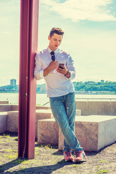 College Student Studying in New York. Wearing white shirt, jeans, sneakers, a young guy standing against pole by Hudson River, looking down, checking messages on his mobile phone. - Photo, image