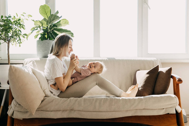 Mãe e bebê sentados em casa em um sofá junto à janela, brincando e rindo. - Foto, Imagem