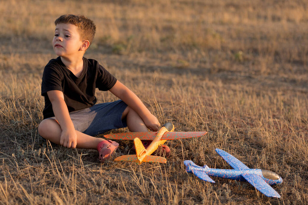zamyślony chłopiec siedzi na suchej trawie, na zewnątrz, w pobliżu leżą samoloty-zabawki, styl życia - Zdjęcie, obraz