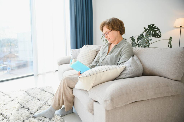 Old lady sitting on couch and enjoying interesting novel stock photo. - 写真・画像
