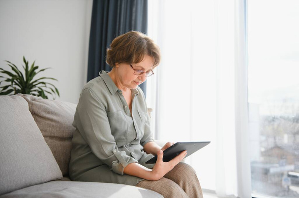 Senior woman looking and laughing at her digital tablet on sofa. - Foto, afbeelding