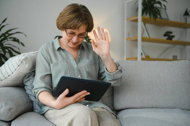 Femme âgée regardant et riant à sa tablette numérique sur le canapé
. - Photo, image