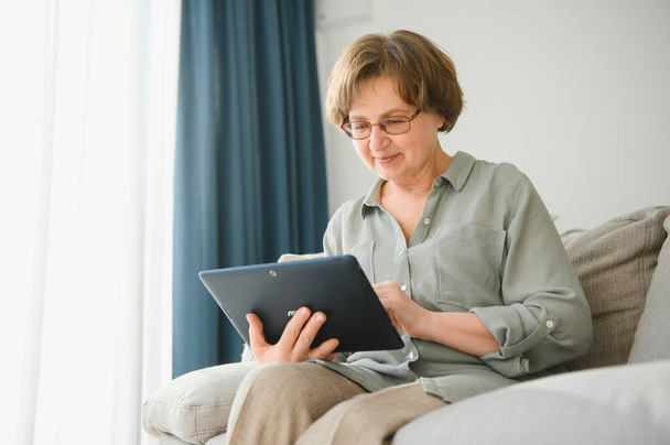 Retirement Leisure. Smiling Elderly Woman Resting With Digital Tablet On Couch At Home, Browsing Internet Or Watching Movies. - Foto, Imagen