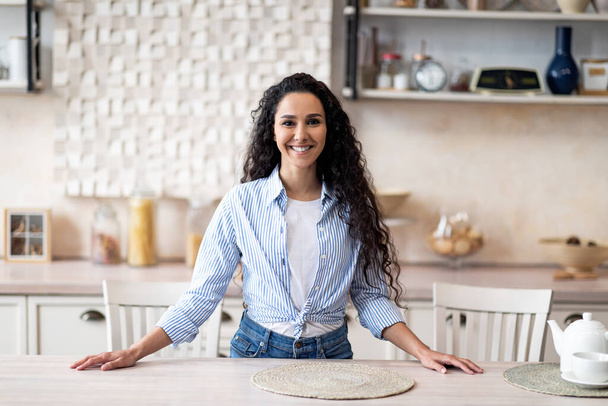 Ritratto di casalinga latina eccitata in piedi vicino al tavolo della cucina, guardando la fotocamera e sorridendo, spazio libero - Foto, immagini