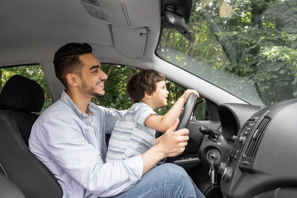 Smiling young man teaching little child to drive car in summer, boy hold steering wheel and look at road, profile - Фото, изображение