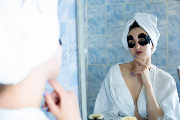 A girl a bathrobe and with a towel on her head sticks patches under her eyes in the bathroom in front of the mirror - Photo, Image