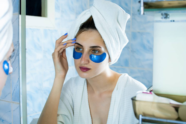 Dziewczyna szlafrok i ręcznik na głowie przykleja łatki pod oczami w łazience przed lustrem. - Zdjęcie, obraz