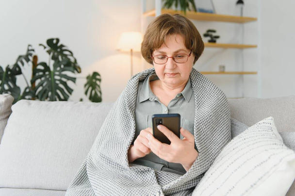 Beautiful senior woman in casual clothing using her smart phone while sitting on the sofa at home. - Foto, Imagem