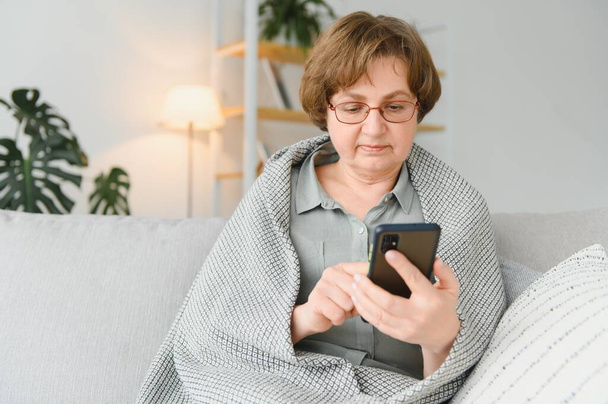 Beautiful senior woman in casual clothing using her smart phone while sitting on the sofa at home. - Фото, изображение