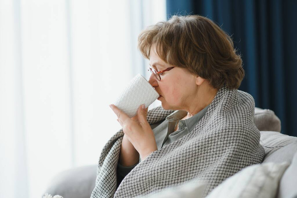 elderly caucasian pensioner wrapped in gray plaid and drinking hot tea in living room. - Foto, Bild