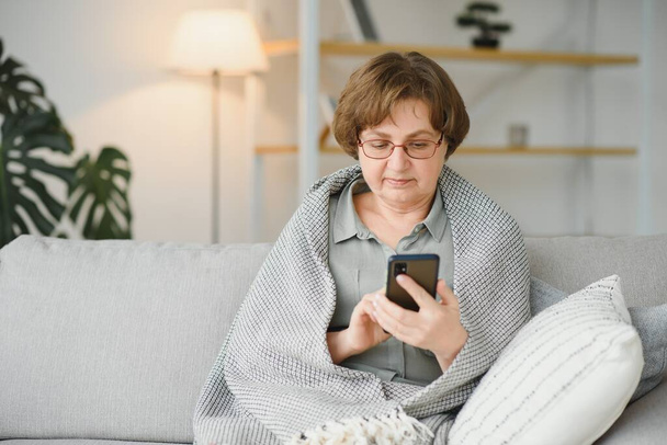 Beautiful senior woman in casual clothing using her smart phone while sitting on the sofa at home. - Photo, Image