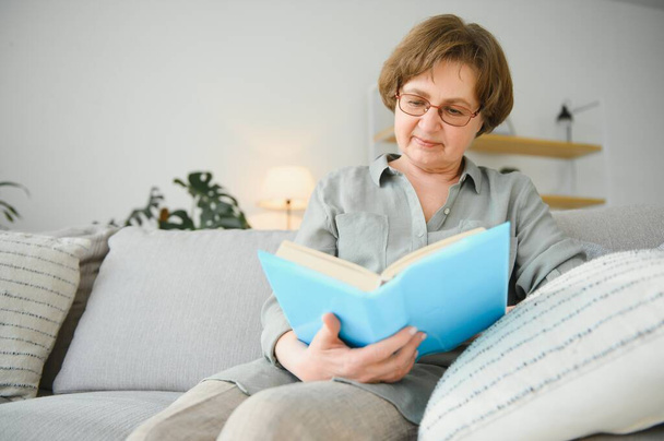 Restful reading. Portrait of thoughtful aged woman reading favorite literature at cozy home. She is lying on pillows on comfortable sofa. Legs are covered with soft blanket - Фото, изображение