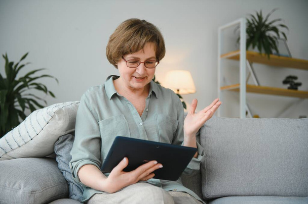 Senior woman looking and laughing at her digital tablet on sofa. - Foto, immagini