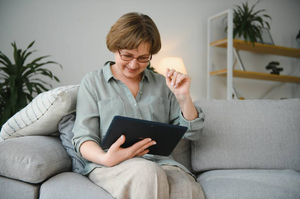 Senior woman looking and laughing at her digital tablet on sofa. - Фото, изображение