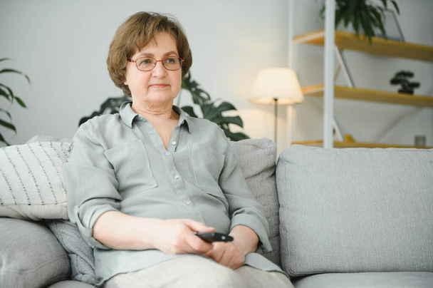people, television and entertainment concept - senior woman in eyeglasses with remote control watching tv at home. - Foto, Bild