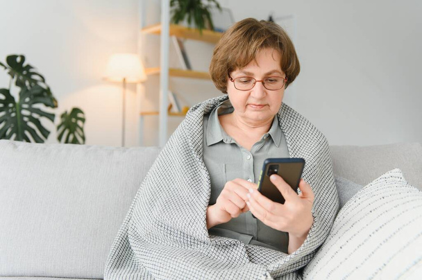 older mature adult woman using smartphone sitting on couch at home. Cheerful grandmother with eyeglasses surfing internet with her mobile phone. Technology concept. - Fotografie, Obrázek