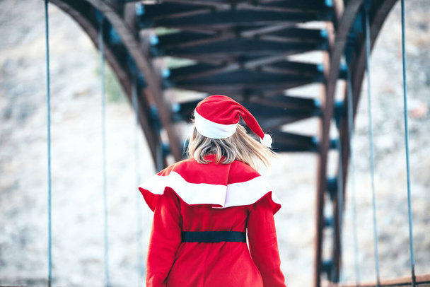 Foto de Mama Noel caminando hacia atrás en un puente de madera con una bolsa roja en la mano - Foto, Imagen
