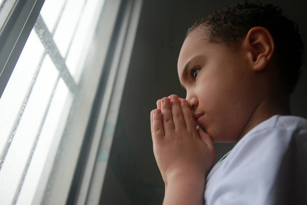 Salvador, Bahia, Brezilya - 20 Nisan 2022: Beyaz etnik kökenli çocuk Salvador şehrinde yağmurlu bir günde bir apartman penceresinin yanında dua ediyor.. - Fotoğraf, Görsel
