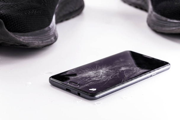 smartphone device fallen on white floor, screen and film cracked in the fall, sneakers in the background, closeup, copy space - 写真・画像