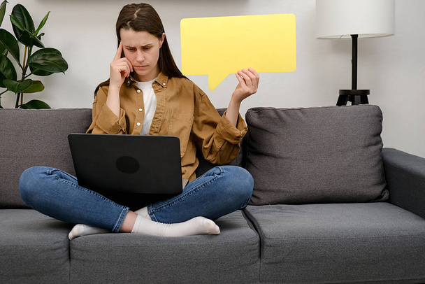 Eine aufgeweckte junge Frau in Hemd sitzt zu Hause auf dem grauen Sofa, der Laptop ist leer, die Sprechblase hält das Kinn in die Höhe. Online-Lernen, Internet-Marketing - Foto, Bild
