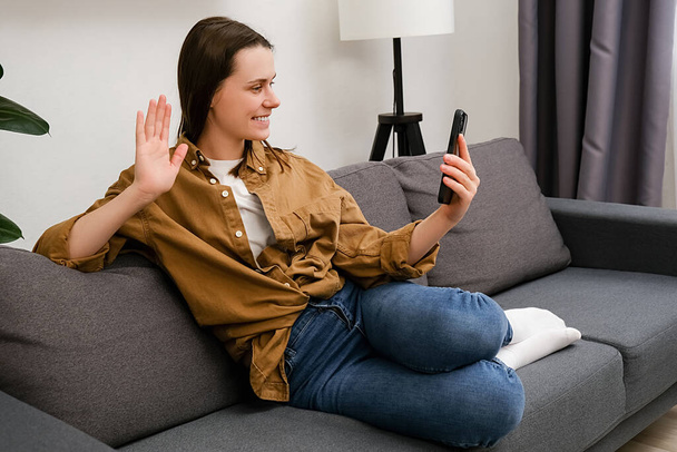 Smiling young female sitting on couch waving hand using smartphone app enjoying online virtual chat video call with friends in distance mobile chat virtual meeting, recording stories for social media - Foto, Imagem