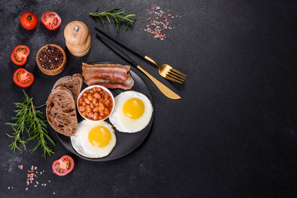 English breakfast with fried eggs, bacon, beans, tomatoes, spices and herbs. A hearty and nutritious start of the day - 写真・画像