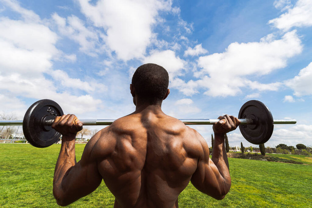 african man with perfect muscular body lifts a barbell with weights in the park -view from behind- - Photo, Image