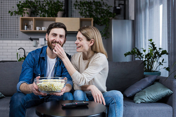 family happy couple man and woman eating popcorn and watching tv sitting on sofa at home - Fotoğraf, Görsel
