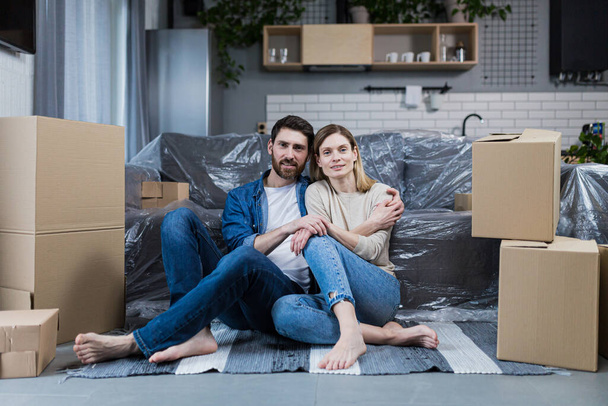 Happy young couple man and woman at home sitting on sofa in new rented apartment, near cardboard boxes - Φωτογραφία, εικόνα
