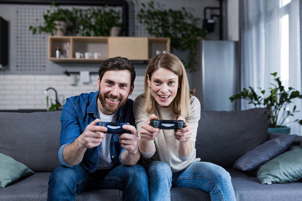 Happy couple man and woman playing game using joystick while sitting on sofa at home - Photo, Image
