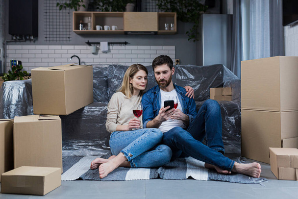 A married couple in love, a man and a woman celebrate a housewarming party, drink wine from cardboard boxes and choose new furniture and household appliances in the online store, use the phone - Foto, Imagem