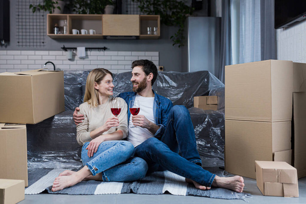 Family couple in love, man and woman sitting on the floor celebrating a new home in a new apartment, drinking wine among cardboard boxes - Φωτογραφία, εικόνα