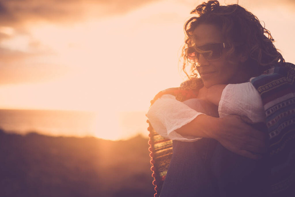 Seitenporträt einer jungen hübschen Frau, die den Sonnenuntergang allein genießt, ihre Knie umarmt und sich selbst liebt. Weibliche Menschen lächeln und entspannen sich in der Abendsonne. Goldene Stunden in der Natur - Foto, Bild