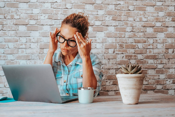 Frau am Schreibtisch am Laptop mit Kopfschmerzen. Konzentrierte und besorgte Geschäftsfrau vor dem Computer. Hausaufgabenplatz mit Mauerziegeln im Hintergrund. Online-Job- und Sicherheitskonzept - Foto, Bild