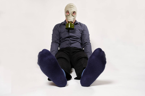 Ein Mann mit Gasmaske sitzt auf weißem Grund, die Beine stinken. Unangenehmer Geruch von Socken bei Männern. - Foto, Bild