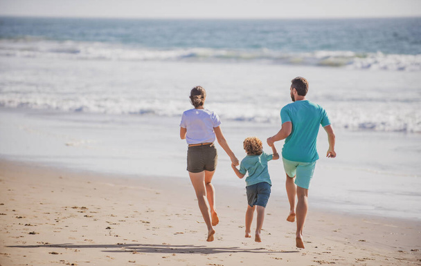 Szczęśliwa młoda rodzina biegnie i skacze po letniej plaży. Koncepcja spędzania wolnego czasu. Dziecko z rodzicami biegającymi i skaczącymi. - Zdjęcie, obraz