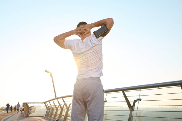 Utrzymuj formę. Widok na umięśnionego sportowca w białej koszulce, noszącego uchwyt na smartfona, rozgrzewającego się o świcie, cieszącego się porannym treningiem przed joggingiem - Zdjęcie, obraz