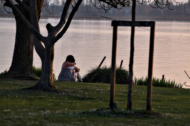 Samotna dziewczyna, która siedzi nad brzegiem jeziora o zachodzie słońca - Zdjęcie, obraz