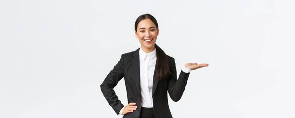 Profesionální usměvavá podnikatelka představí svůj projekt během setkání. Prodejkyně v černém obleku drží ruku vpravo jako ukazující produkt, drží na dlani přes prázdné bílé pozadí - Fotografie, Obrázek