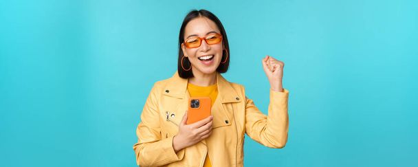 Захоплена усміхнена азіатська дівчина в сонцезахисних окулярах, тримає мобільний телефон і танцює, сміється зі смартфона, стоїть на синьому фоні
 - Фото, зображення