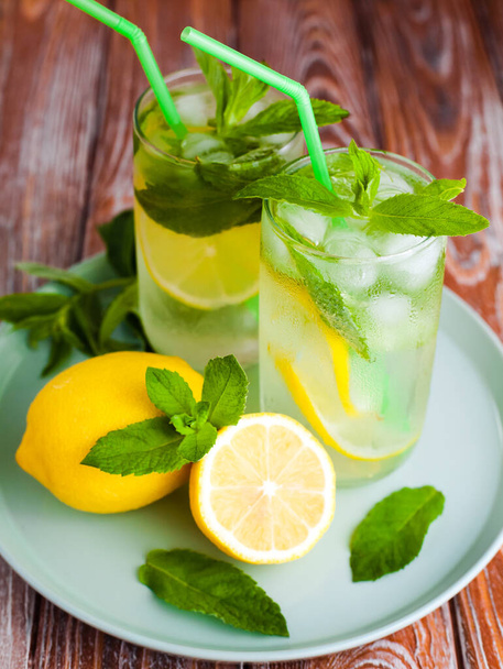 Two glass with natural refreshing lemonade or mojito cocktail with lemon, mint and ice cubes. Cold summer beverage on wooden background. Close-up. - Foto, Imagem