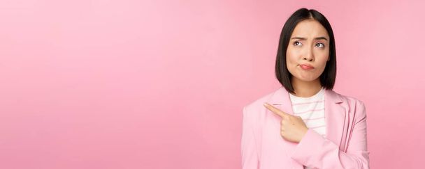 Donna d'affari dubbia, bronza e smorfia mentre indica, guardando a sinistra a banner con espressione scettica del viso, in posa su sfondo rosa - Foto, immagini