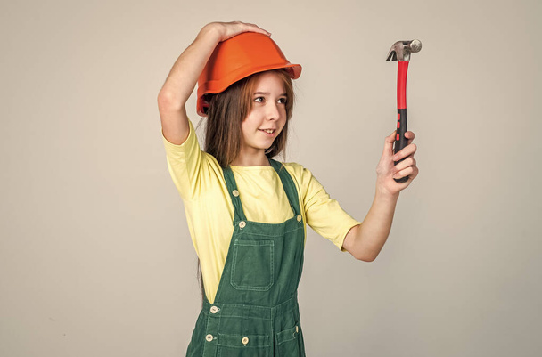 teen girl laborer in protective helmet and uniform on grey background use hammer, worker - Фото, изображение