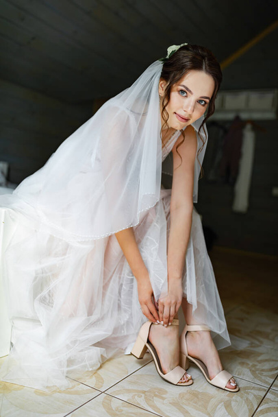 Fiatal barna menyasszony esküvői ruhában pózol az ablak mellett. Leejtett vállheveder. - Fotó, kép