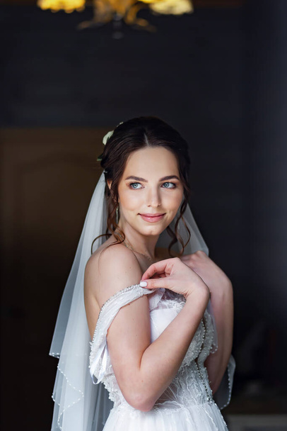 Young brunette bride in a wedding dress posing near the window. Drop shoulder strap. - 写真・画像