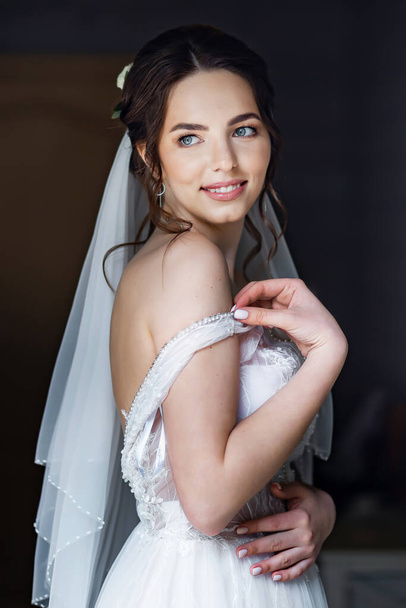 Fiatal barna menyasszony esküvői ruhában pózol az ablak mellett. Leejtett vállheveder. - Fotó, kép